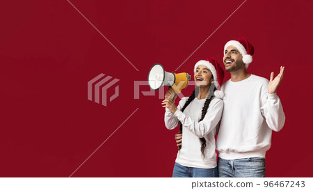 Arab Couple In Santa Hats Making Announcement With Loudspeaker Over Red Background Arab Couple In Santa Hats Making Announcement With Loudspeaker Over Red Background 96467243