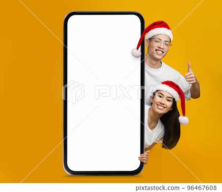 Happy chinese young woman and guy in Santa Claus hats show thumb up with big smartphone with blank screen Happy chinese young woman and guy in Santa Claus hats show thumb up with big smartphone with blank screen 96467608