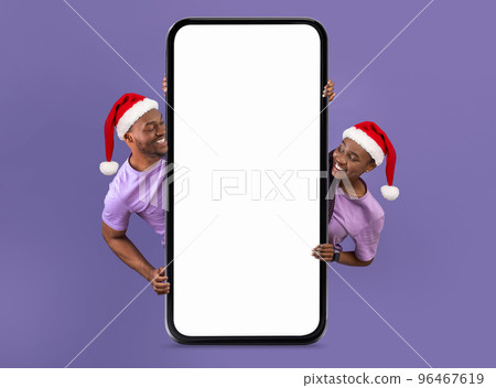 Cheerful young black woman and man in Christmas hats looking from behind huge phone with blank screen 96467619