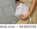 Closeup shot of female hands holding beautiful Christmas present with pink ribbon on white background in interior 96468348