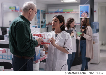 Senior client holding cardiology pills package asking for pharmacist medical advice during medication shopping in drugstore. Helpful druggist selling medicinal supplements. Health care service 96469437