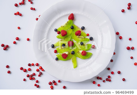 christmas food for kids. white dish with christmas tree made of kiwi, raspberries, cowberry and huckleberry 96469499