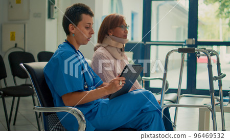Nurse consulting patient with neck collar brace and walking frame, taking notes about accident injury on tablet. Doing checkup visit with woman wearing cervical foam and impairment. Nurse consulting patient with neck collar brace and walking frame, taking notes about accident injury on tablet. Doing checkup visit with woman wearing cervical foam and impairment. 96469532