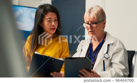 Elderly medic and patient looking at digital tablet and report papers, filling in medical checkup form to attend consultation appointment. Doctor analyzing documents in clinic lobby. Handheld shot. 96469700