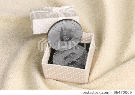 The silver bitcoin lies in a small orange gift box with a small bow on a blanket made of soft and fluffy light orange fleece fabric with a large number of relief folds The silver bitcoin lies in a small orange gift box with a small bow on a blanket made of soft and fluffy light orange fleece fabric with a large number of relief folds 96470069