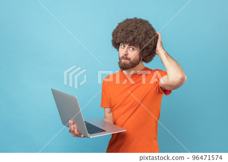 Portrait of man with Afro hairstyle wearing orange T-shirt holding laptop and thinking about new project, scratching the back of his head. Indoor studio shot isolated on blue background. 96471574