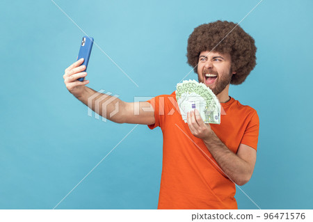 Portrait of excited crazy man with Afro hairstyle wearing orange T-shirt having video call or livestream, showing euro banknotes to subscribers. Indoor studio shot isolated on blue background. 96471576