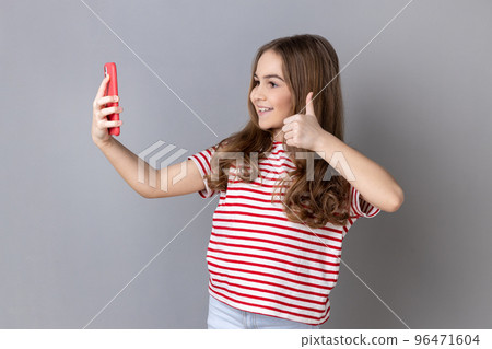 Portrait of little girl wearing striped T-shirt making video call using smartphone, smiling with toothy smile, showing thumb up. Indoor studio shot isolated on gray background. 96471604