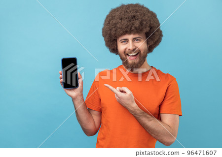 Portrait of smiling satisfied man with Afro hairstyle wearing orange T-shirt pointing at mobile phone with finger, showing empty space for advertisement. Indoor studio shot isolated on blue background 96471607
