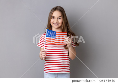 American flag. Portrait of delighted happy adorable cute little girl wearing striped T-shirt holding flag of United States of America, looking at camera. Indoor studio shot isolated on gray background 96471610