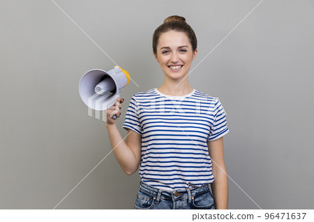 Portrait of woman wearing striped T-shirt having satisfied expression, holding megaphone in hands, looking at camera with toothy smile. Indoor studio shot isolated on gray background. 96471637