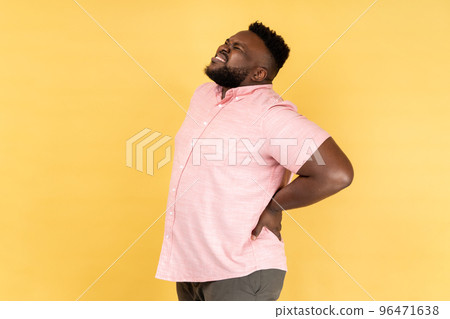 Back, kidney or spine pain. Portrait of man wearing pink shirt standing and endure pain on back, frowning face, suffering terrible pain. Indoor studio shot isolated on yellow background. Back, kidney or spine pain. Portrait of man wearing pink shirt standing and endure pain on back, frowning face, suffering terrible pain. Indoor studio shot isolated on yellow background. 96471638