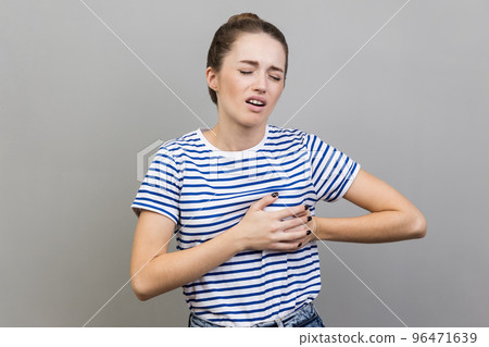 Acute pain in chest. Portrait of woman in striped T-shirt clutching breast and grimacing from painful cramp, heart attack at young age, cardiac disease. Indoor studio shot isolated on gray background. Acute pain in chest. Portrait of woman in striped T-shirt clutching breast and grimacing from painful cramp, heart attack at young age, cardiac disease. Indoor studio shot isolated on gray background. 96471639