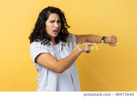 Portrait of confused woman with dark wavy hair pointing wrist watch and looking annoyed displeased, showing clock to hurry up. Indoor studio shot isolated on yellow background. Portrait of confused woman with dark wavy hair pointing wrist watch and looking annoyed displeased, showing clock to hurry up. Indoor studio shot isolated on yellow background. 96471640