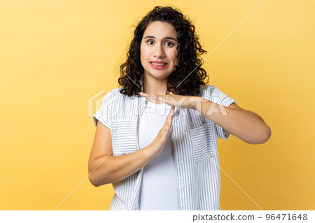 Portrait of bossy serious woman with dark wavy hair showing time out gesture, looking with strict expression at camera, deadline. Indoor studio shot isolated on yellow background. 96471648