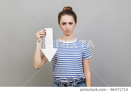 Portrait of sad upset woman wearing striped T-shirt showing white arrow pointing down, expressing sadness and sorrow, downgrade concept. Indoor studio shot isolated on gray background. 96471719