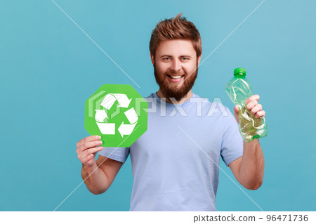 Portrait of smiling optimistic handsome bearded man pressing bottle holding in hand recycling sign board, plastic disposal, garbage sorting. Indoor studio shot isolated on blue background. Portrait of smiling optimistic handsome bearded man pressing bottle holding in hand recycling sign board, plastic disposal, garbage sorting. Indoor studio shot isolated on blue background. 96471736
