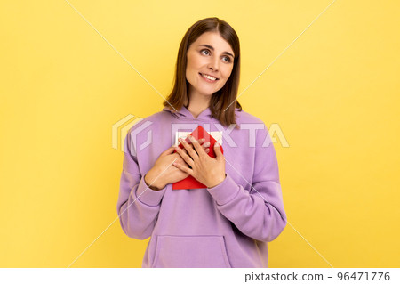 Portrait of beautiful woman standing, embracing envelope and looking away with pleasant smile and enjoying, wearing purple hoodie. Indoor studio shot isolated on yellow background. Portrait of beautiful woman standing, embracing envelope and looking away with pleasant smile and enjoying, wearing purple hoodie. Indoor studio shot isolated on yellow background. 96471776