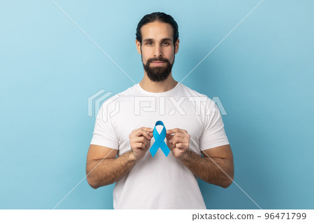 Portrait of serious responsible man with beard wearing white T-shirt holding blue awareness, disease symbol, looking at camera, support. Indoor studio shot isolated on blue background. 96471799