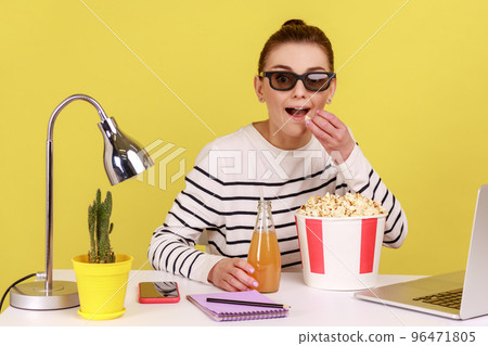 Excited satisfied woman in 3d glasses eating popcorn with beverage, sitting on workplace and enjoying break, enjoying interesting film. Indoor studio studio shot isolated on yellow background. Excited satisfied woman in 3d glasses eating popcorn with beverage, sitting on workplace and enjoying break, enjoying interesting film. Indoor studio studio shot isolated on yellow background. 96471805