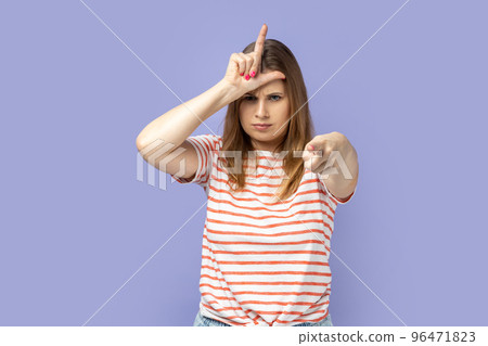 You are loser. Portrait of serious blond woman wearing striped T-shirt showing loser gesture and pointing finger on you, abuser. Indoor studio shot isolated on purple background. 96471823