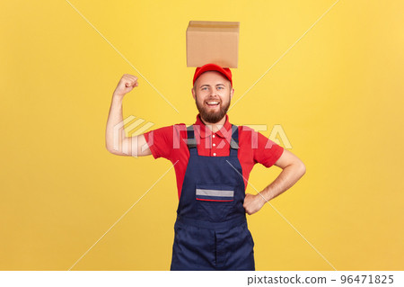 Portrait of strong courier man wearing blue uniform standing with cardboard box on his head, looking at camera, raised arm showing his power. Indoor studio shot isolated on yellow background. 96471825