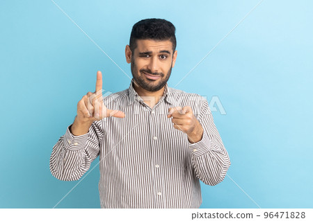 Portrait of disrespectful rude young businessman showing loser gesture and pointing on you, abuser, frowning face, wearing striped shirt. Indoor studio shot isolated on blue background. 96471828