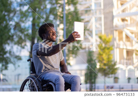 Young guy in wheelchair having video chat on tablet outdoors 96473133