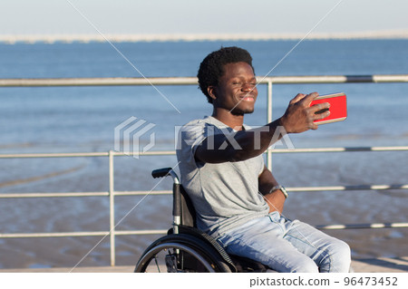 Young guy in wheelchair taking selfie against sea background 96473452