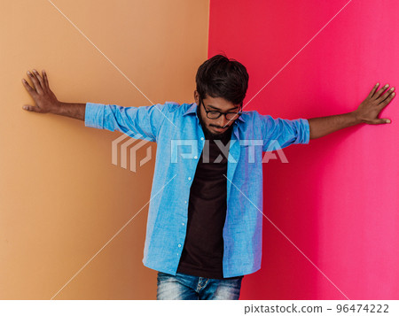 A young Indian student in a blue shirt with glasses posing on pink-orange background  96474222