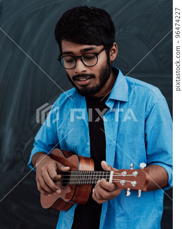 Indian young man in a blue shirt and glasses playing the guitar in front of the school blackboard Indian young man in a blue shirt and glasses playing the guitar in front of the school blackboard 96474227