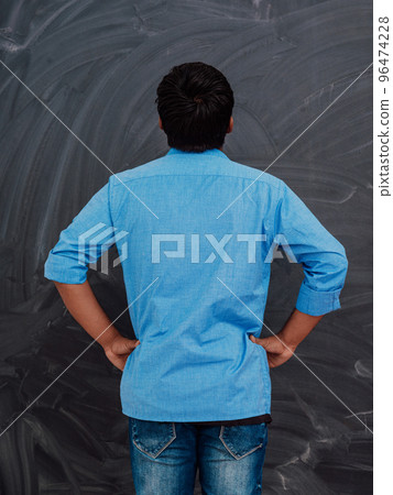 A young Indian student in a blue shirt with his back to the camera is standing in front of the school blackboard A young Indian student in a blue shirt with his back to the camera is standing in front of the school blackboard 96474228