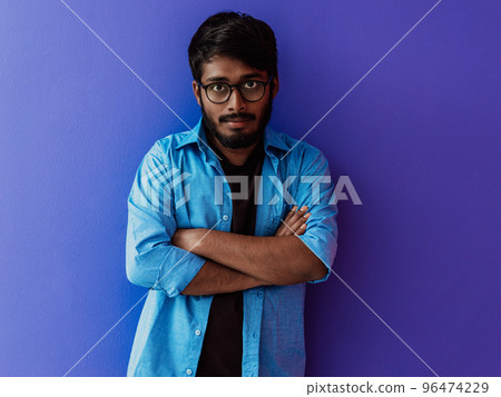 A young Indian student in a blue shirt with glasses posing with his arms crossed posing on purple  96474229