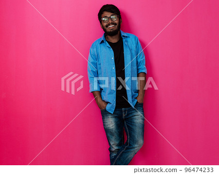 Indian smiling young man with blue shirt and glasses posing on pink background Indian smiling young man with blue shirt and glasses posing on pink background 96474233