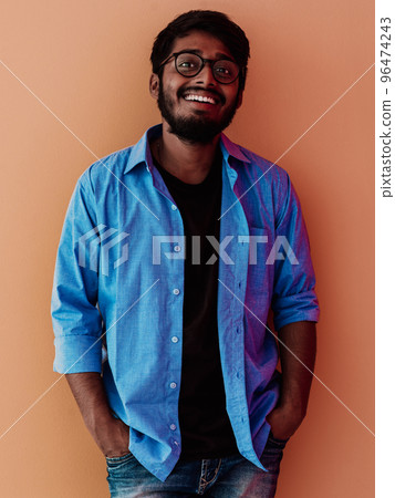 Indian smiling young student in blue shirt and glasses posing on orange background Indian smiling young student in blue shirt and glasses posing on orange background 96474243