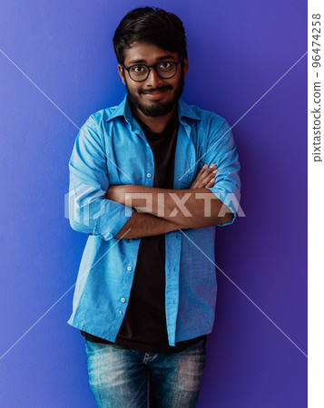 A young Indian student in a blue shirt with glasses posing with his arms crossed posing on purple  96474258