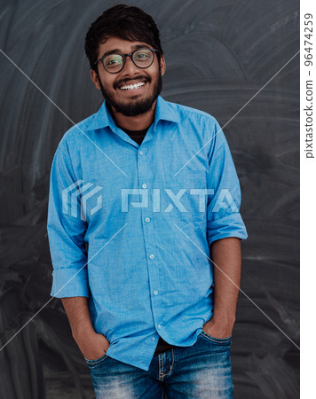 Indian smiling young student in blue shirt and glasses posing on school blackboard background Indian smiling young student in blue shirt and glasses posing on school blackboard background 96474259