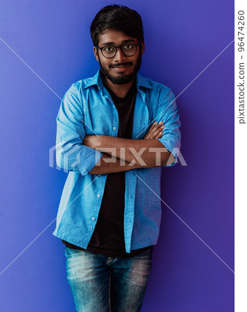 A young Indian student in a blue shirt with glasses posing with his arms crossed posing on purple 96474260