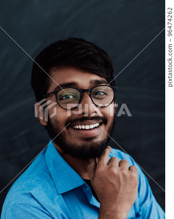 A young Indian man in a blue shirt and glasses poses thoughtfully in front of school board A young Indian man in a blue shirt and glasses poses thoughtfully in front of school board 96474262