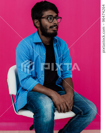 A young Indian student in a blue shirt with glasses sitting in front of a pink background A young Indian student in a blue shirt with glasses sitting in front of a pink background 96474264
