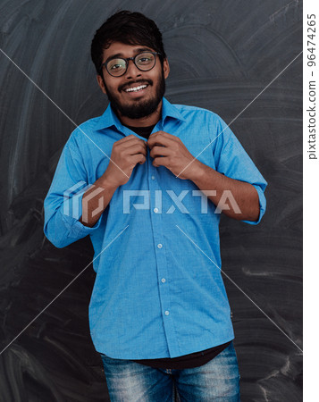 Indian young student wearing glasses puts on a blue shirt while standing in front of the school blackboard Indian young student wearing glasses puts on a blue shirt while standing in front of the school blackboard 96474265
