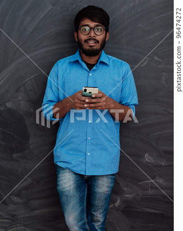Indian smiling young student in blue shirt and glasses using smartphone and posing on school blackboard background 96474272
