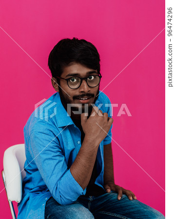 A young Indian student in a blue shirt with glasses sitting in front of a pink background A young Indian student in a blue shirt with glasses sitting in front of a pink background 96474296