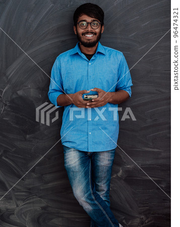 Indian smiling young student in blue shirt and glasses using smartphone and posing on school blackboard background Indian smiling young student in blue shirt and glasses using smartphone and posing on school blackboard background 96474321