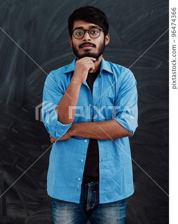 A young Indian man in a blue shirt and glasses poses thoughtfully in front of school board A young Indian man in a blue shirt and glasses poses thoughtfully in front of school board 96474366