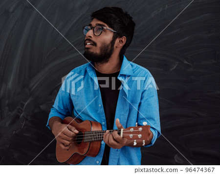 Indian young man in a blue shirt and glasses playing the guitar in front of the school blackboard Indian young man in a blue shirt and glasses playing the guitar in front of the school blackboard 96474367