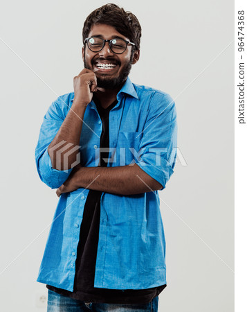 A young Indian man in a blue shirt and glasses poses thoughtfully in front of a gray background A young Indian man in a blue shirt and glasses poses thoughtfully in front of a gray background 96474368