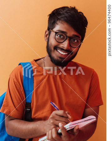 Indian student with blue backpack, glasses and notebook posing on orange background. The concept of education and schooling. Time to go back to school Indian student with blue backpack, glasses and notebook posing on orange background. The concept of education and schooling. Time to go back to school 96474400