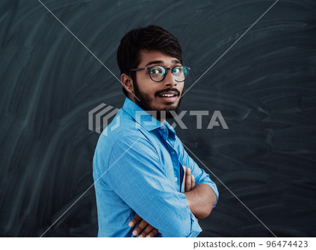 A young Indian student in a blue shirt with glasses posing with his arms crossed in front of the school blackboard 96474423