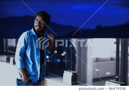 An Indian man with glasses and a blue shirt looks around the city at night. In the background of the night street of the city 96474596